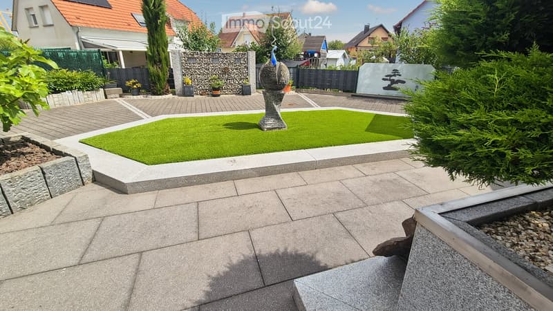 Well-maintained garden with artificial grass, stone pathways, decorative stone sculpture, and surrounding greenery.