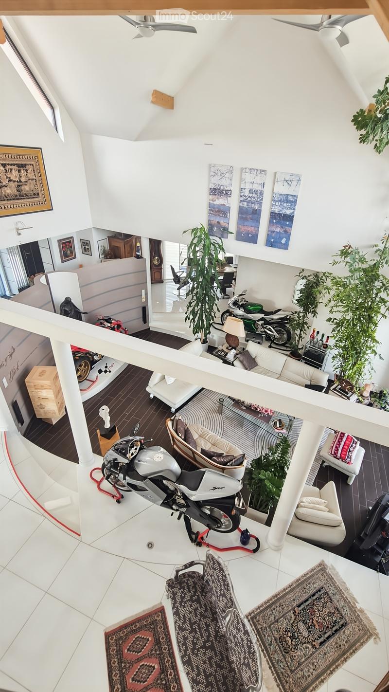 Open-plan living space with white walls, wooden floors, and modern decor. Features include a white sofa, a chair, a small table with a lamp, a motorcycle on a stand, several potted plants, and artwork on the walls.