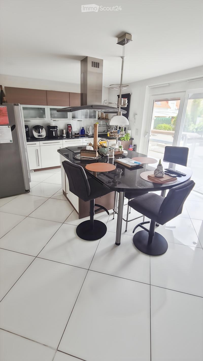 Modern kitchen with white tiled flooring, white cabinets, and stainless steel appliances. Features a central island with a black countertop, a round dining table with four chairs, and various kitchen appliances including a refrigerator, coffee maker, and