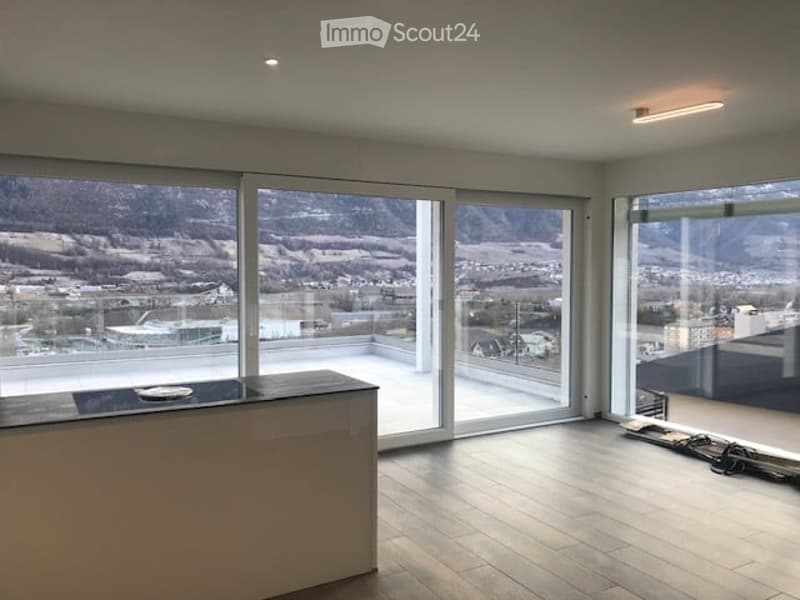 Bright, empty room with large glass sliding doors leading to a balcony, offering a scenic mountain view. The room features a modern design with wooden flooring and minimalistic decor.