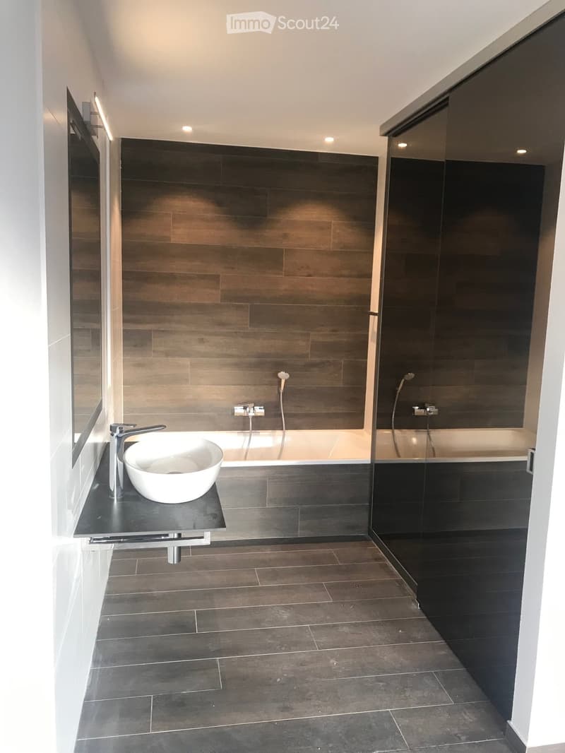 Modern bathroom with dark tiled walls and floor, a white sink bowl on a black countertop, a bathtub with a glass shower enclosure, and sleek fixtures.