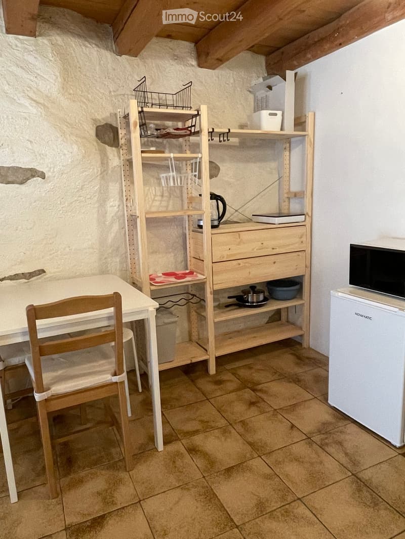 A small, cozy kitchen area with a table, chair, shelves, a refrigerator, microwave, and various kitchen items.