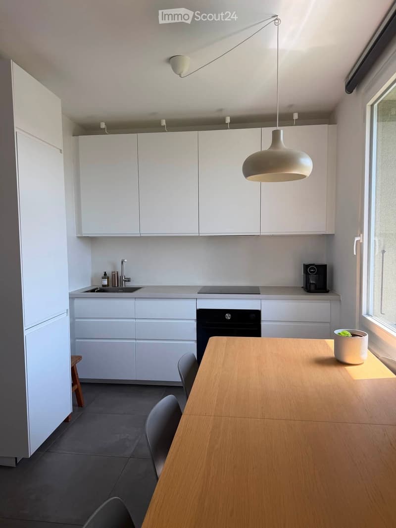 Modern kitchen with white cabinets, a wooden dining table, grey chairs, and a pendant light. Features a sink, faucet, oven, and coffee maker on the counter.