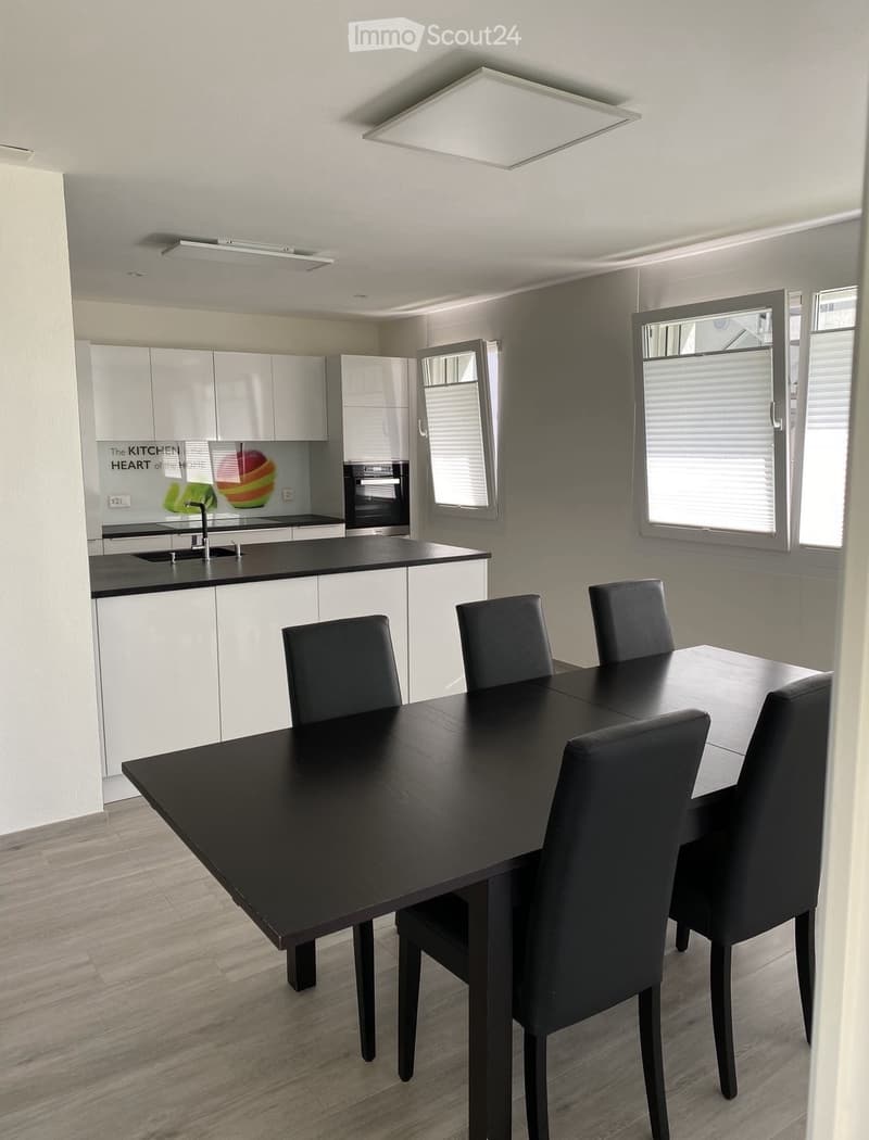 A modern dining room with a large black dining table and chairs, white kitchen cabinets, a kitchen area with an oven and a sink, white tiled flooring, and large windows with white blinds.
