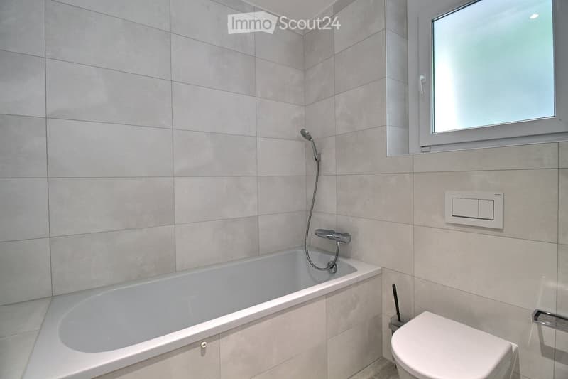 Modern bathroom with a bathtub, toilet, showerhead, and a window with frosted glass. The walls are tiled in light gray, and there is a control panel for the shower on the wall.