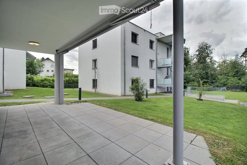 Modern outdoor patio area with tiled flooring, surrounded by a grassy lawn and a white multi-story building with balconies and windows.