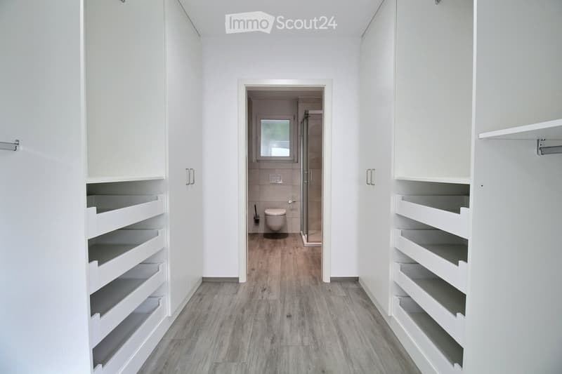 A clean, modern hallway with white built-in storage units on both sides, leading to a small bathroom with a toilet, shower, and mirror.