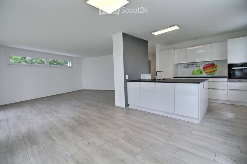 Modern, minimalist kitchen with white cabinets, a central island, and a sleek black countertop. The space features light wood flooring and large windows allowing natural light to fill the room.