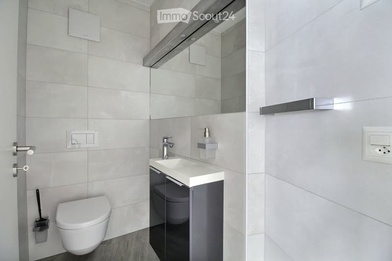 Modern bathroom with white tiles, a toilet, a sink with a modern faucet, a large mirror with built-in lighting, and a soap dispenser. The walls are tiled in light grey, and the floor is dark wood.