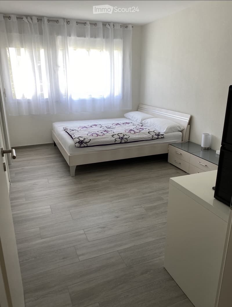 Modern bedroom with a single bed, nightstand, lamp, and drawer unit. Light wooden flooring and white walls.