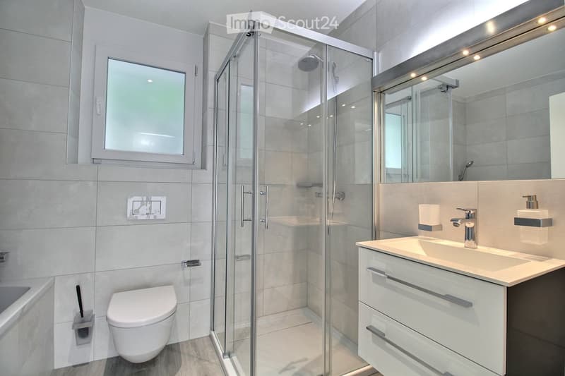 Modern bathroom with a glass-enclosed shower, toilet, sink, and bathtub. Features include a large mirror, sleek fixtures, and a window for natural light.