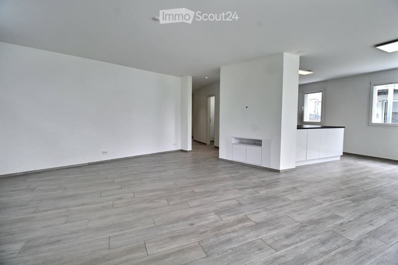 Empty room with light grey walls, wooden flooring, and white cabinetry along one wall. Two windows are visible, and the space is well-lit with recessed lighting.