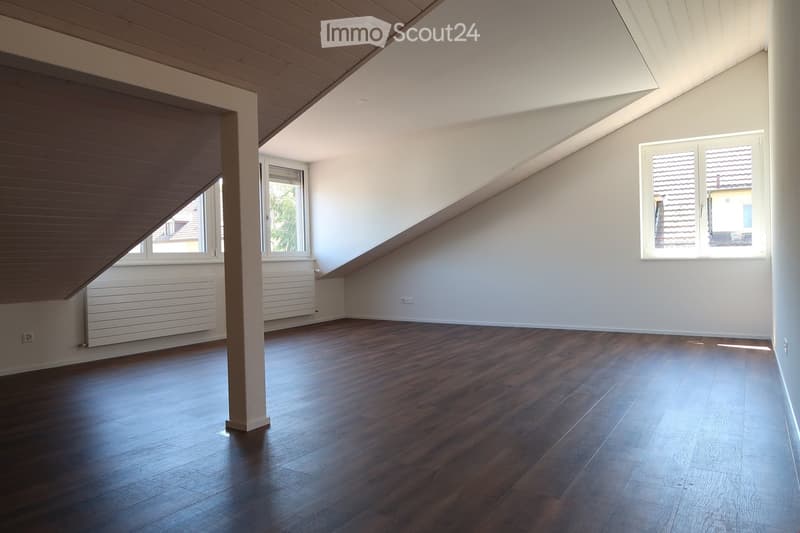 Empty attic room with wooden flooring, white walls, and large windows allowing natural light. The room features a slanted ceiling and a central support column.