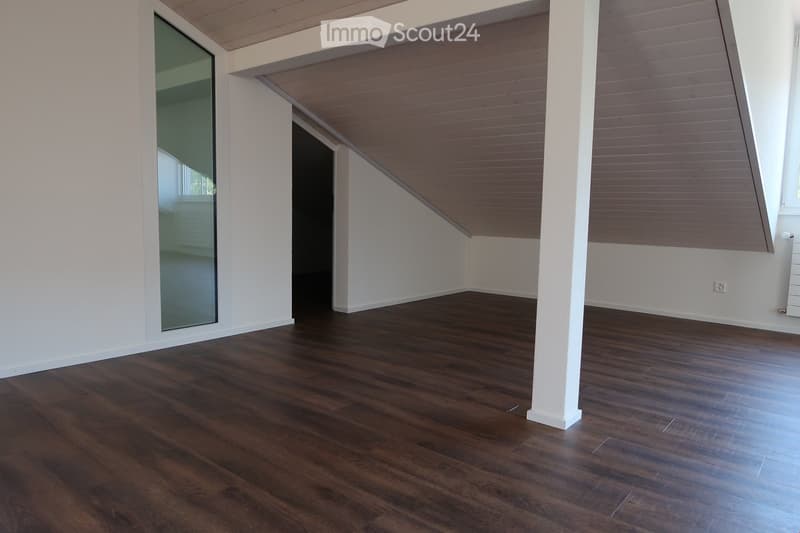 Empty room with wooden flooring, white walls, and a slanted wooden ceiling. Features a large window and a door leading to another space.
