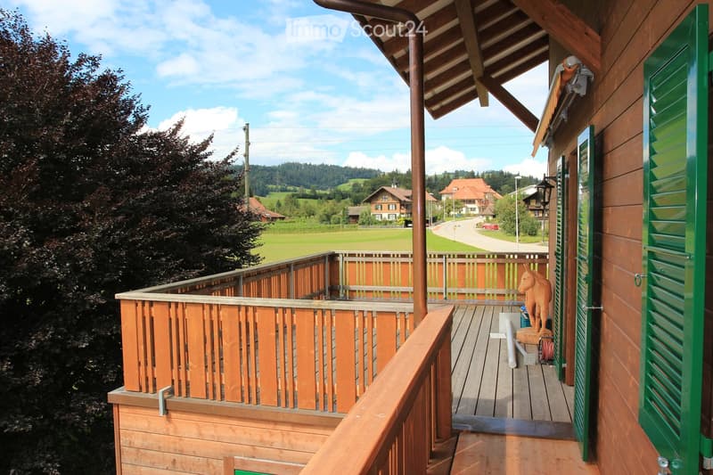 Wooden balcony with railing, overlooking a scenic countryside with houses and green fields.