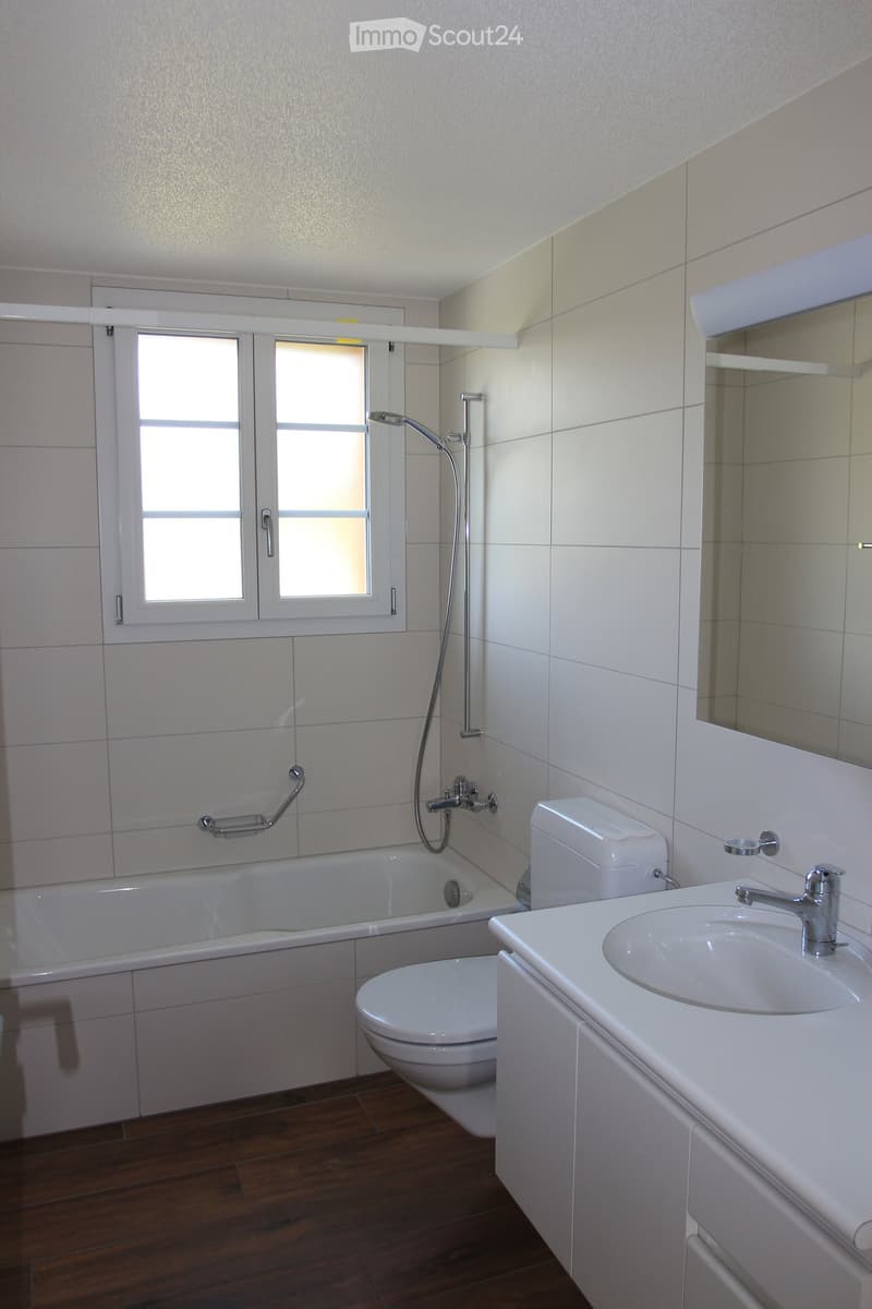 Modern bathroom with white tiles, a bathtub, toilet, sink, and a showerhead. Features a window with frosted glass panes and a wooden floor.