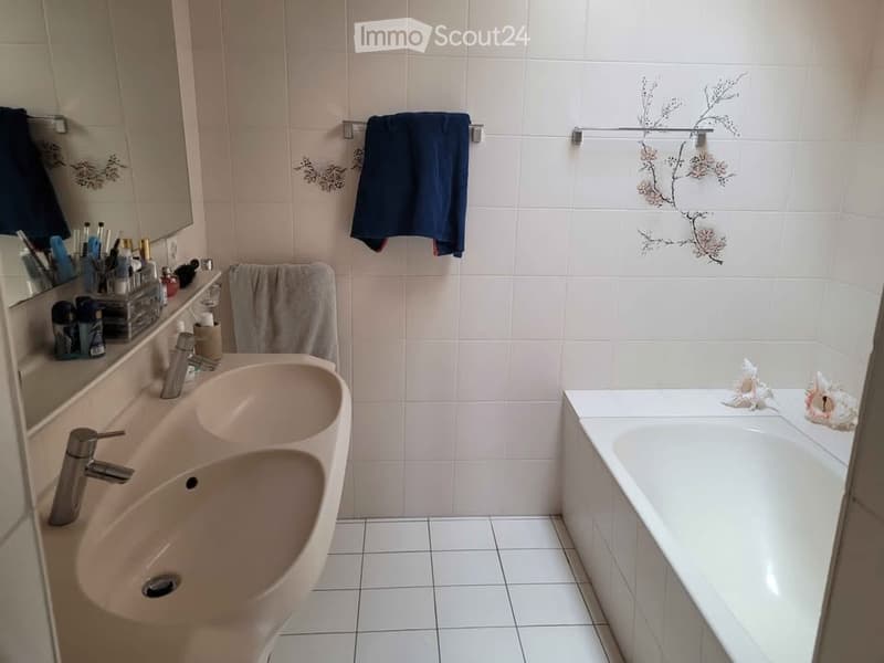 A modern bathroom with white tiled walls and floor, featuring a double sink, a bathtub, a mirror, and decorative towels.