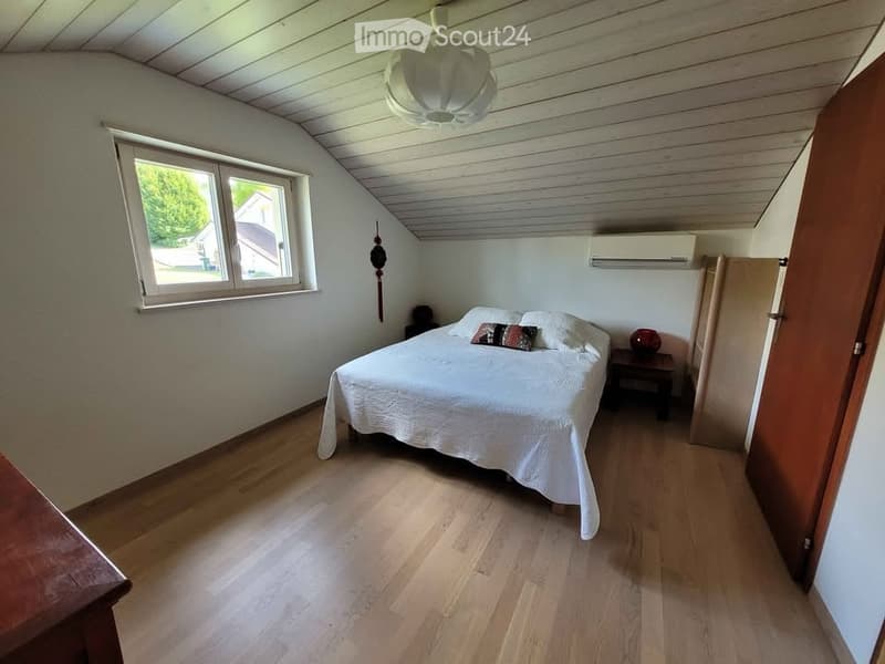 Cozy attic bedroom with wooden floors, slanted ceiling, large window, bed with white bedding, nightstand, and decorative items.
