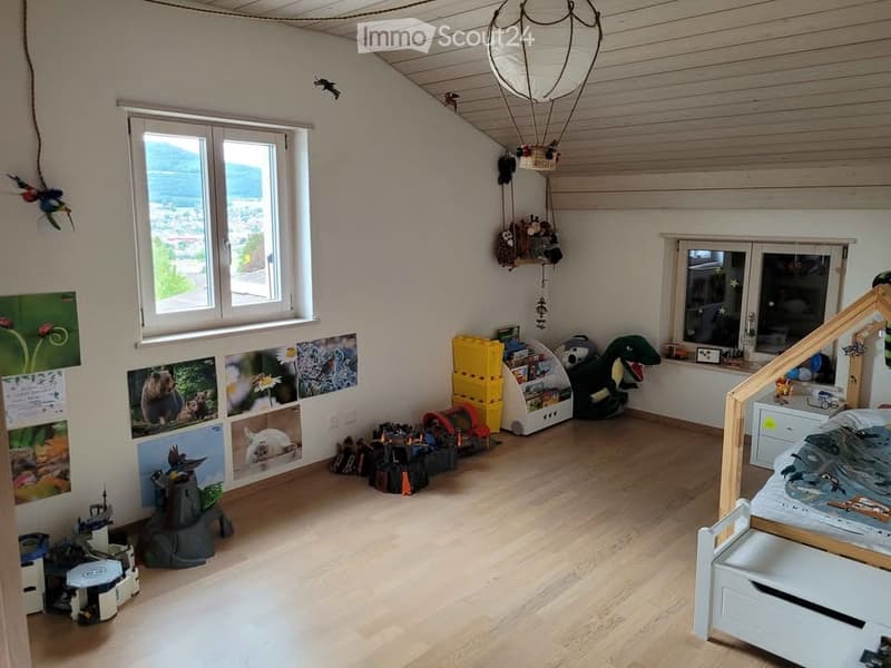 Bright, airy children's room with wooden flooring, large windows offering a scenic view, a bed with a themed bedspread, a colorful toy box, a bookshelf, a plush toy dinosaur, and various toys and decorations including picture frames and a balloon decorati