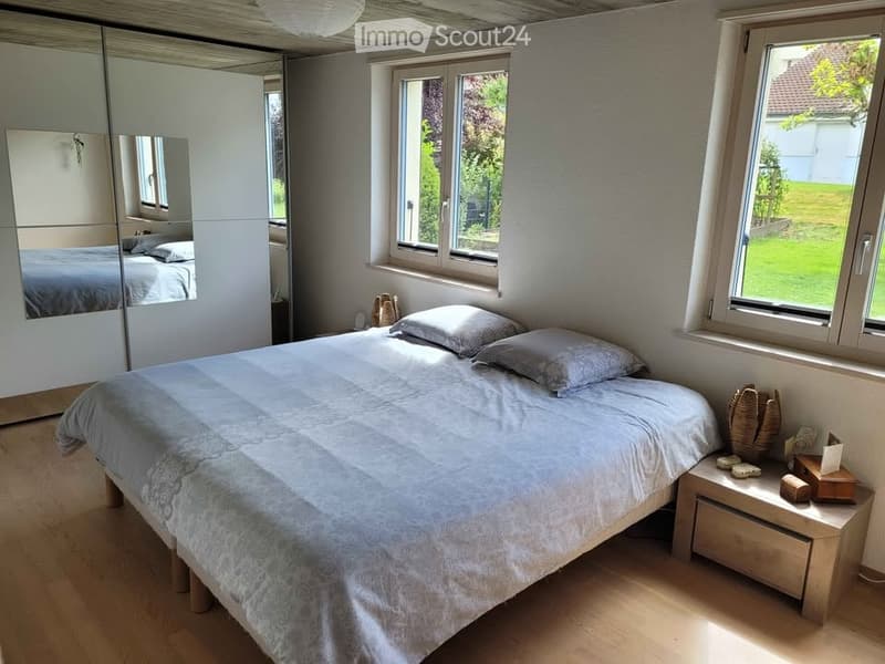 Bright bedroom with a double bed, nightstand, mirror, and wardrobe. Large windows provide natural light and a view of the garden.