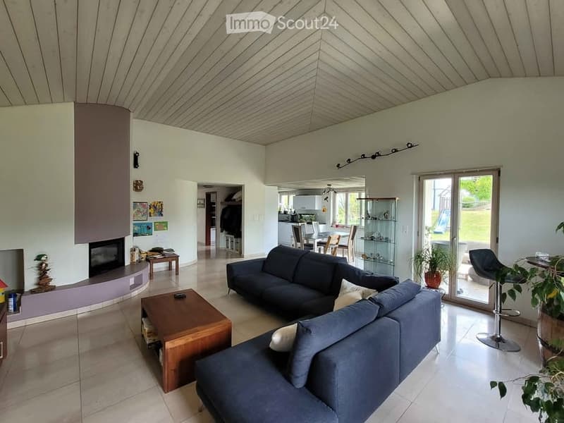 Modern living room with a fireplace, two blue sofas, a wooden coffee table, and a small stool. The room features large windows and a view of the garden.