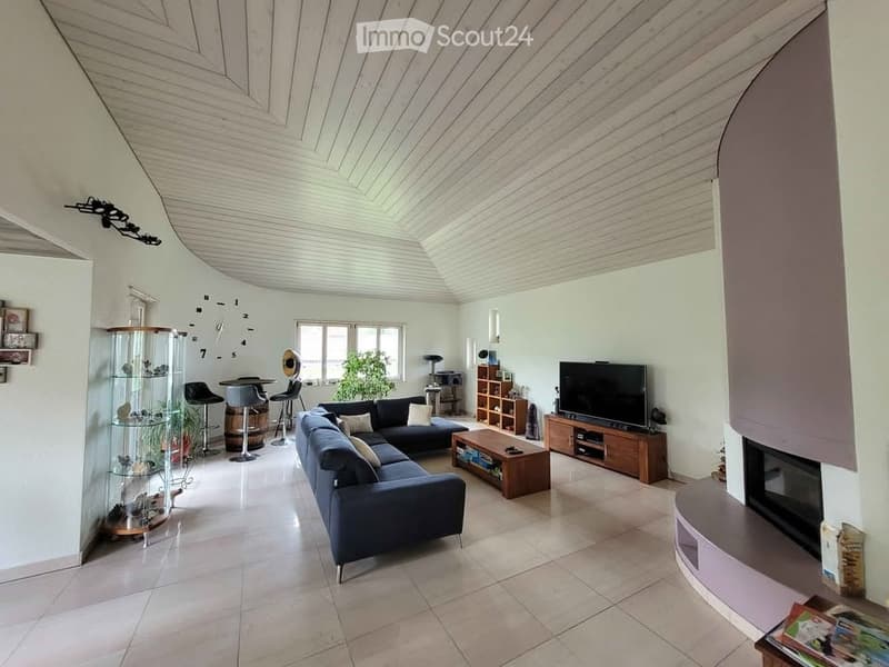 Bright living room with a modern design, featuring a sectional sofa, coffee table, fireplace, and large windows. The space includes a bar area with stools and a display cabinet.
