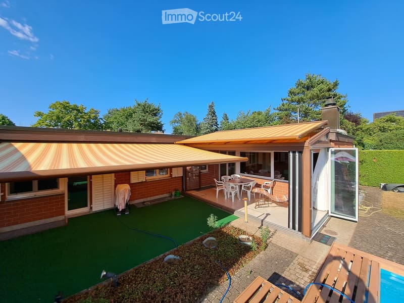 A modern house with a spacious outdoor area featuring a covered patio, dining set, lounge chairs, artificial grass, small garden beds, and a swimming pool.