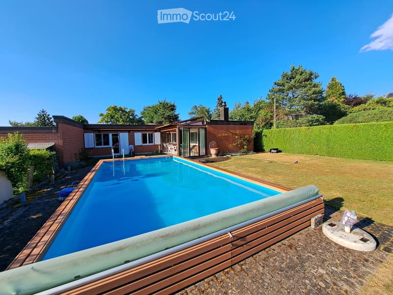 Outdoor area with a swimming pool, wooden decking, surrounding lawn, and a brick house with large windows and shutters in the background.