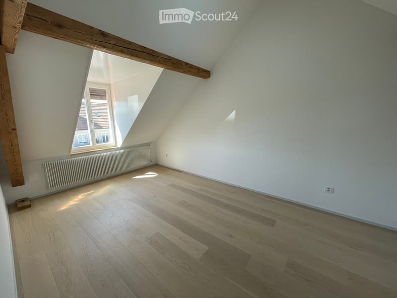 Unfurnished Room. Empty attic room with wooden flooring, white walls, exposed wooden beams, and a window letting in natural light.