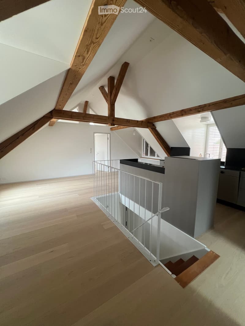 Bright, open attic corridor with wooden beams, light wooden flooring, and a modern metal railing leading to a staircase.