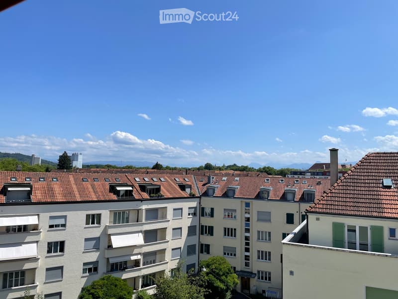 Multi-story residential building with red-tiled roofs, white walls, and multiple windows, surrounded by trees and distant mountains.