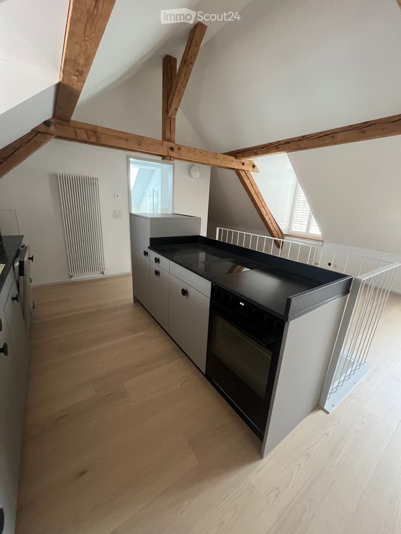 Modern kitchen with light wooden flooring, grey cabinets, black countertop, and built-in oven. Exposed wooden beams on the ceiling and large windows provide natural light.