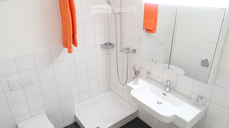 Modern bathroom with white tiles, a shower, a sink with a mirror, and an orange towel hanging on the wall.