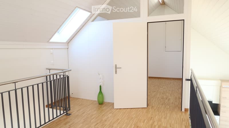 Bright, modern hallway with wooden flooring, white walls, and a skylight. Features a green vase with white flowers on the floor and a closed door leading to another room.