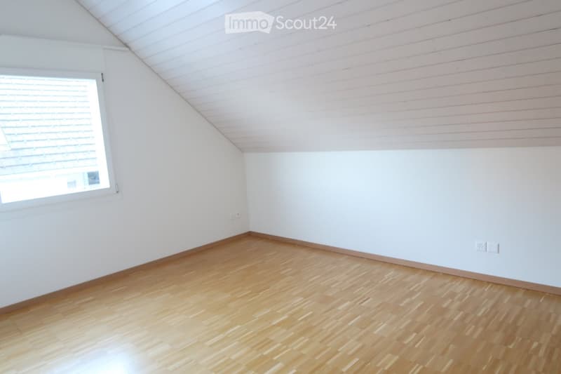 Empty room with wooden flooring, white walls, and a slanted ceiling. A single window with blinds is visible on one wall.