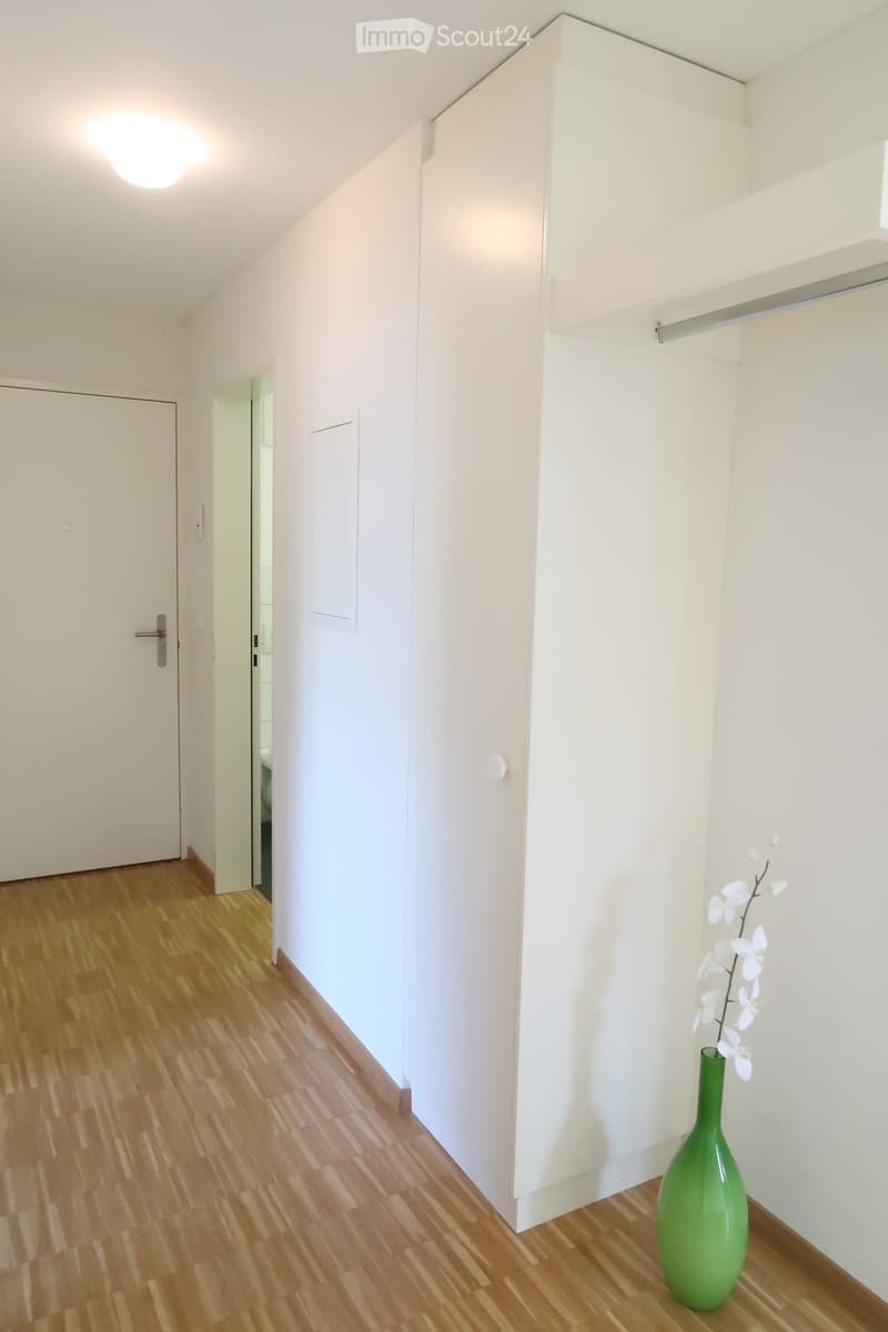 A clean, minimalist hallway with light wooden flooring, white walls, and a single green vase with white flowers on the right side. The hallway features a closed door on the left and an open doorway leading to another room.