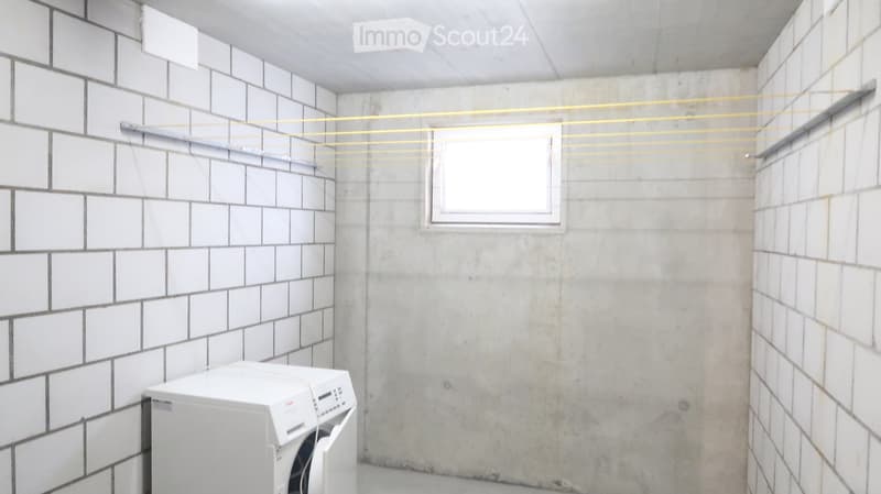 A small, unfinished room with concrete walls and a tiled section, featuring a washing machine and a clothesline with multiple hanging lines.