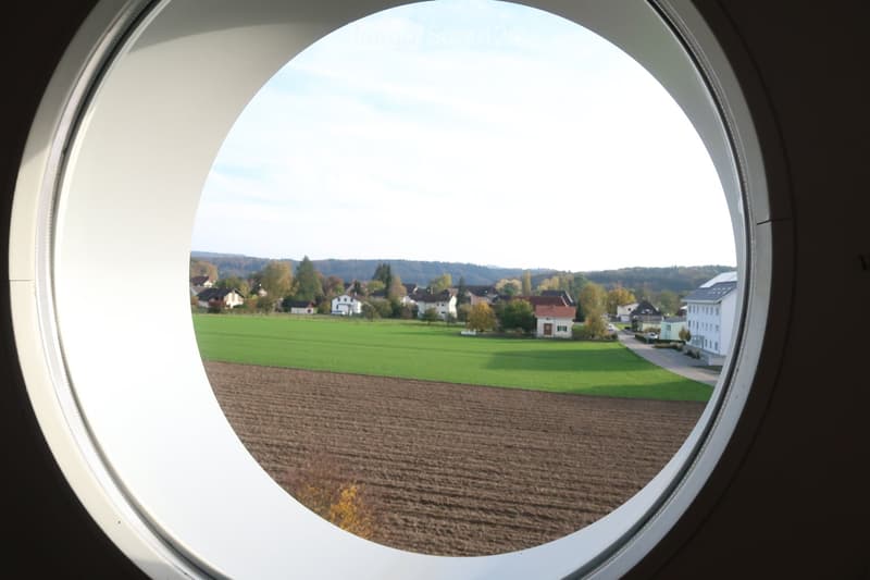 A view through a large, round window showing a rural landscape with fields, houses, and trees.