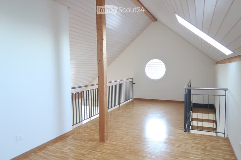 Bright, open hallway with wooden flooring, white walls, and a slanted wooden ceiling. Features a staircase with metal railings and a round skylight window providing natural light.