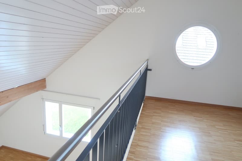 Modern hallway with wooden flooring, white walls, and a metal railing leading to an upper level. Features a round window and a door with glass panels.