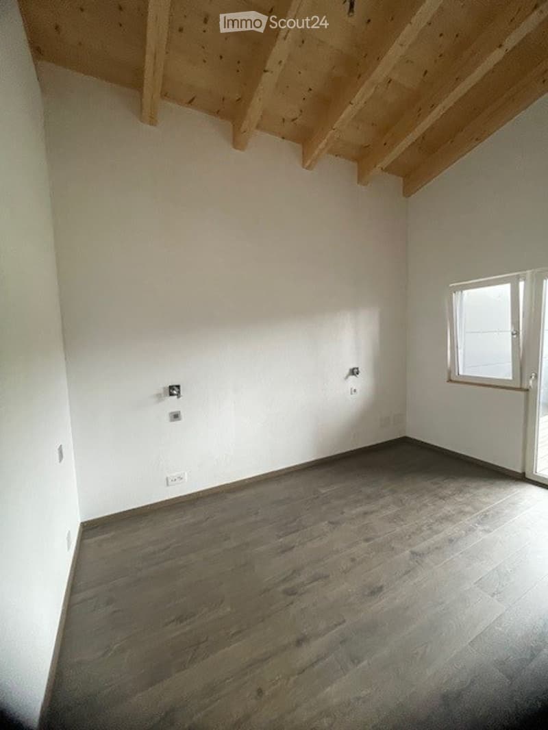 Empty room with wooden floor and white walls, featuring exposed wooden beams on the ceiling and a single window with white frame.