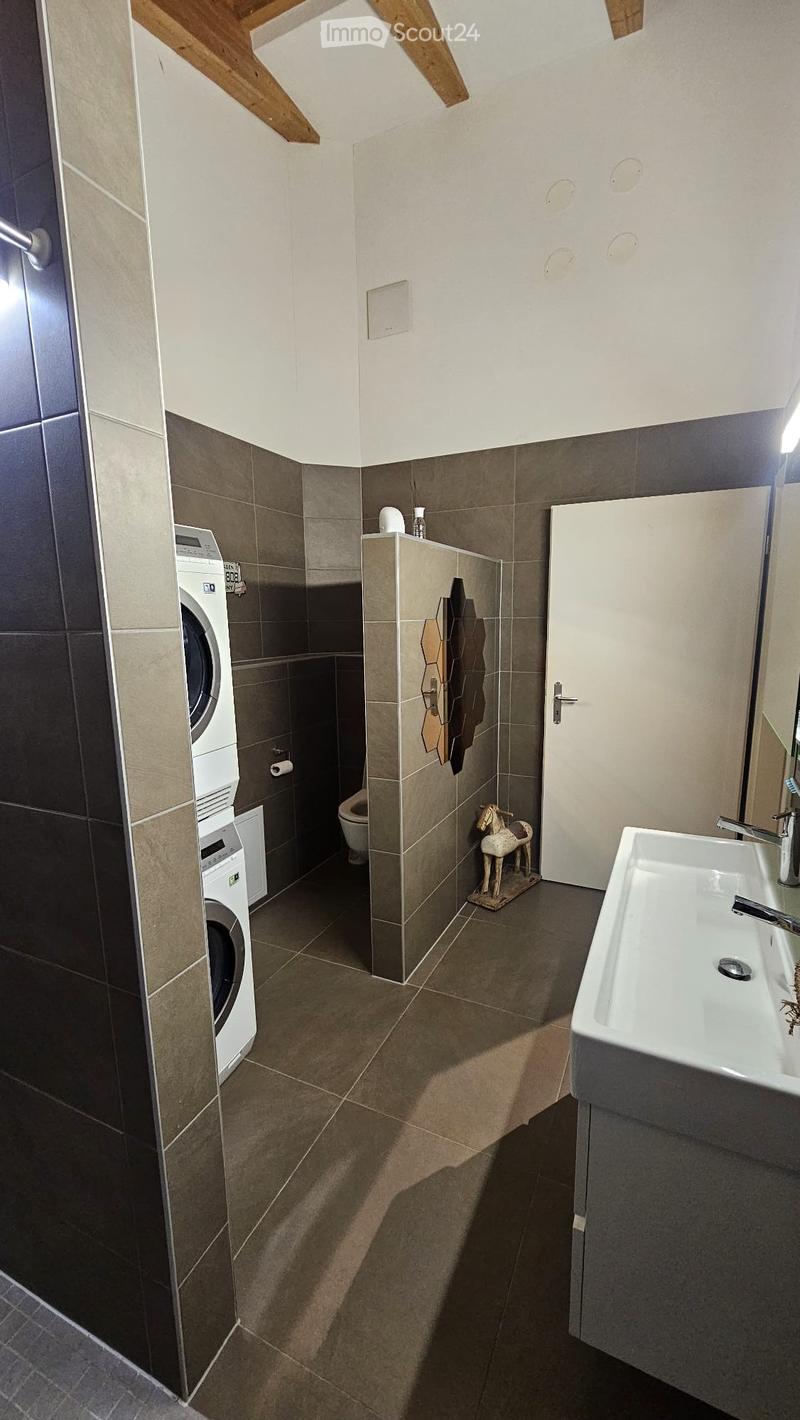 Modern bathroom with a white sink, washing machine, toilet, and decorative mirror. Features a sleek, contemporary design with dark tiles and a wooden accent wall.