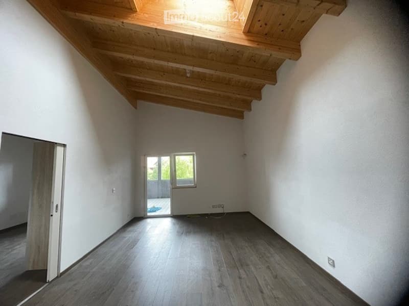 Empty room with wooden flooring, white walls, and exposed wooden ceiling beams. A doorway leads to an outdoor area with a visible view of greenery.