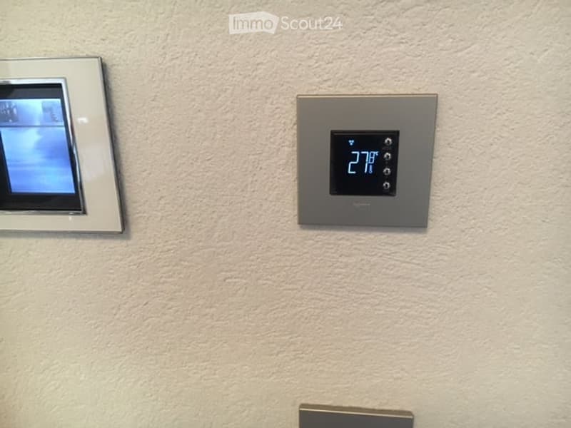 Empty room with a textured beige wall, featuring a digital thermostat displaying 27°F and a small screen or display device mounted on the wall.