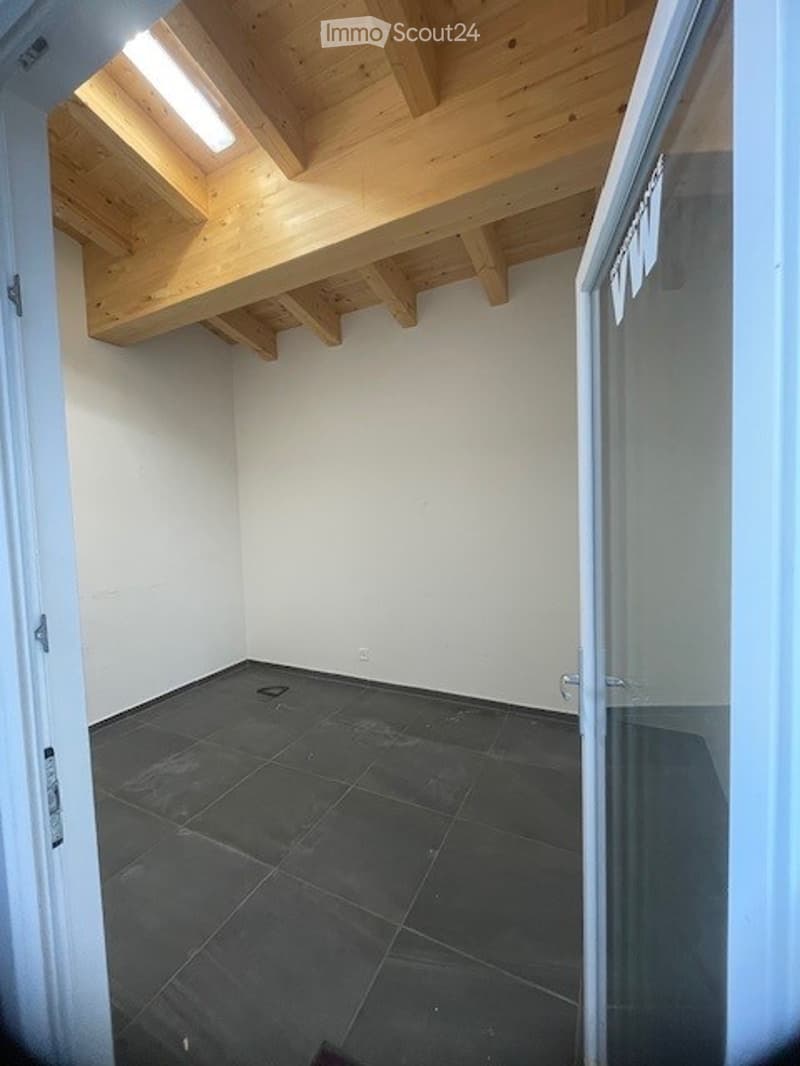 Empty room with exposed wooden beams on the ceiling, dark tiled floor, white walls, and a glass door leading outside.