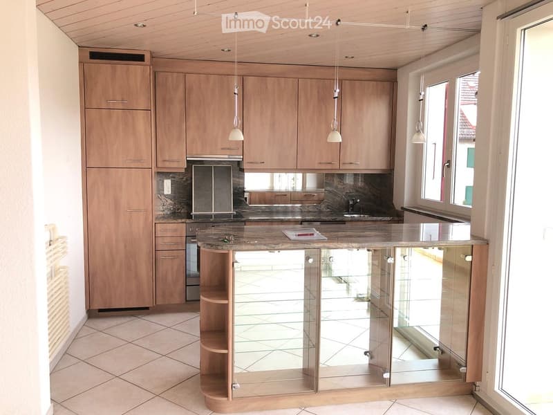 Modern kitchen with light wooden cabinets, a central island with glass panels, and pendant lights hanging from the ceiling.