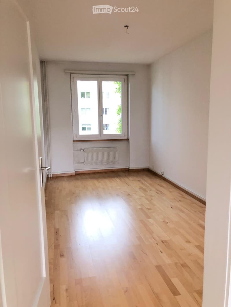Empty room with light wooden flooring, white walls, and a large window with double doors leading outside.