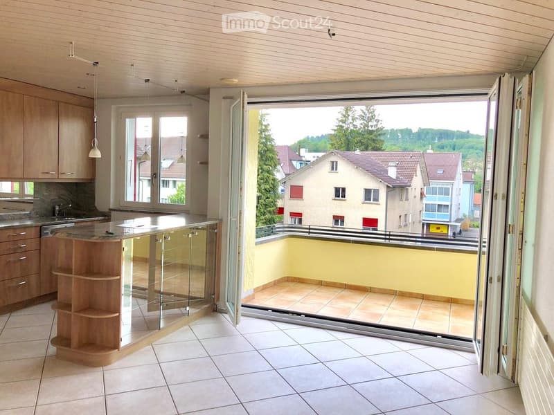Modern kitchen with light wood cabinets, a central island with glass railing, and pendant lights. The space opens to a balcony with a view of the neighborhood.