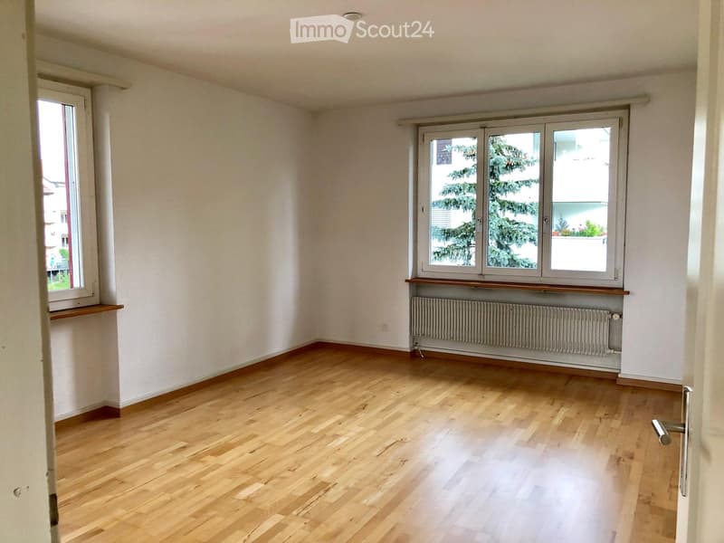 Empty room with light wooden flooring, white walls, and large windows with wooden sills. A radiator is visible under one window.
