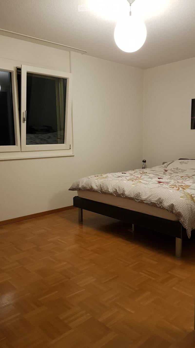 A modern bedroom with wooden flooring, a single bed with a patterned bedspread, and a large window with white frames and curtains.